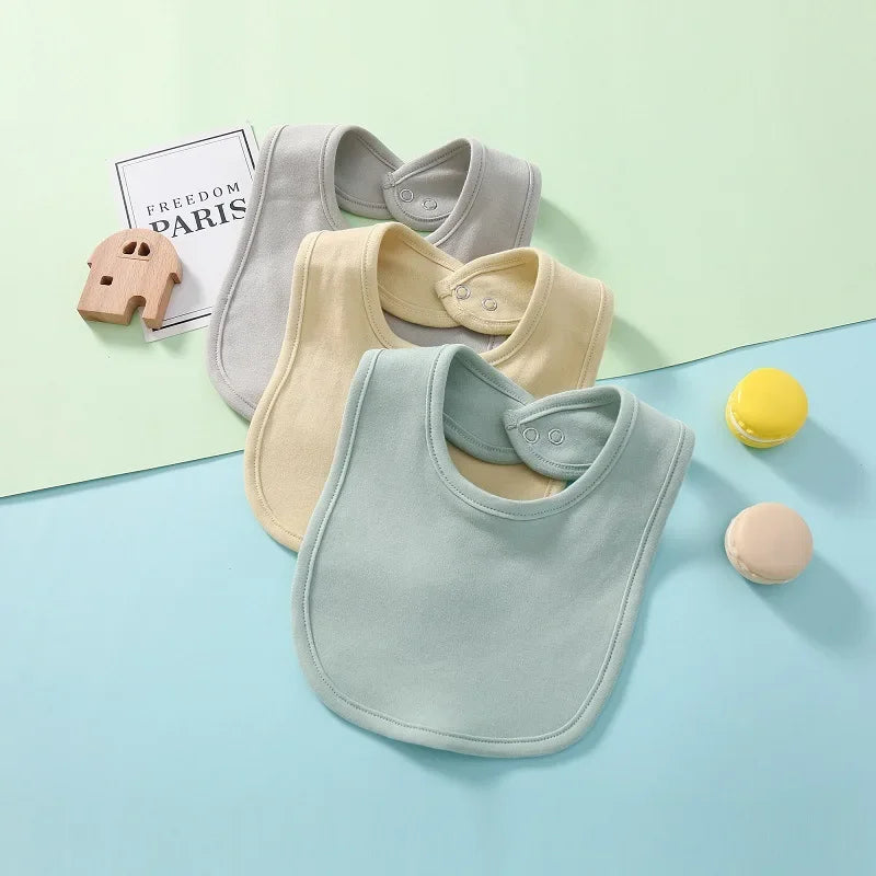 Three baby bibs in gray, beige, and light blue on a pastel background with toys.