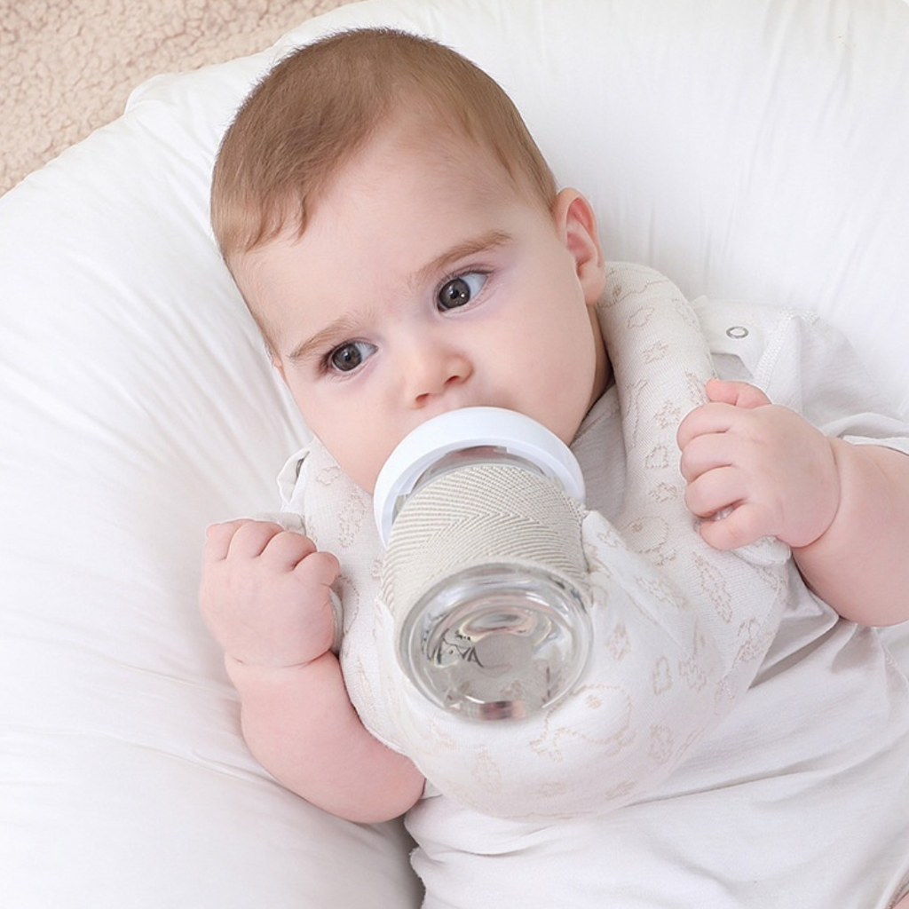 Baby holding a bottle pillow, lying on a white pillow.