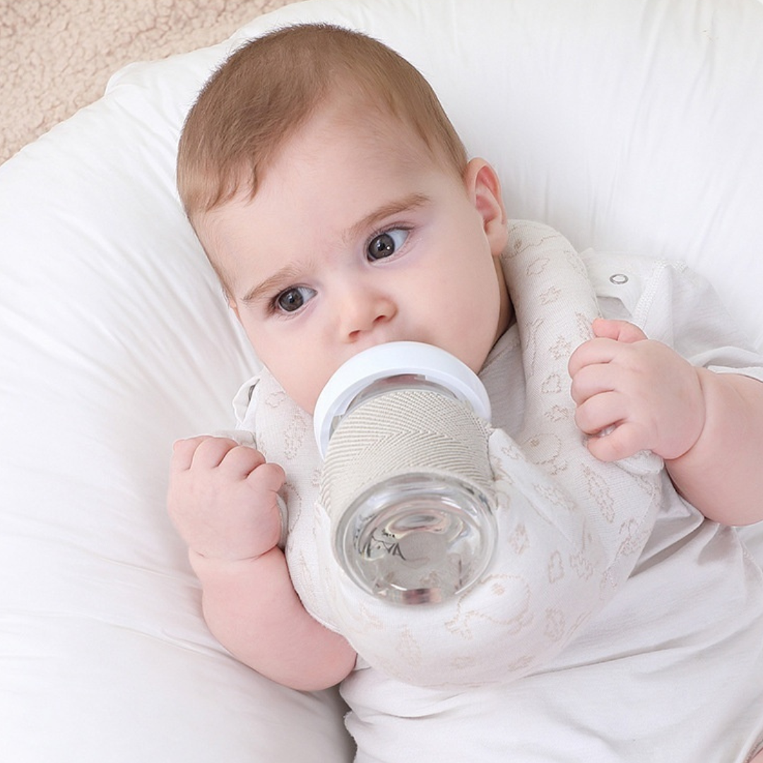 Baby holding a bottle pillow, lying on a white pillow.